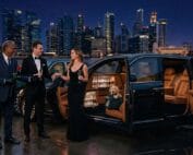 A couple in formal evening wear sharing champagne beside an open luxury sedan with a chauffeur presenting a rose, city skyline lit up at night in the background