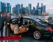 A chauffeur and driver assisting a woman with a small dog and luggage into a black luxury sedan, red carpet rolled out, city skyline and harbor in the background at dusk