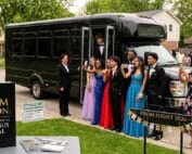 Prom students in formal wear posing for photos in front of a black party bus with a chauffeur and parents nearby, "Prom Season: A Parent's Guide to Party Bus Rental" sign displayed