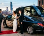 A bride and groom holding hands beside a chauffeur-driven luxury sedan and Sprinter shuttle van in downtown Chicago at dusk, with a chauffeur assisting and the Willis Tower skyline in the background